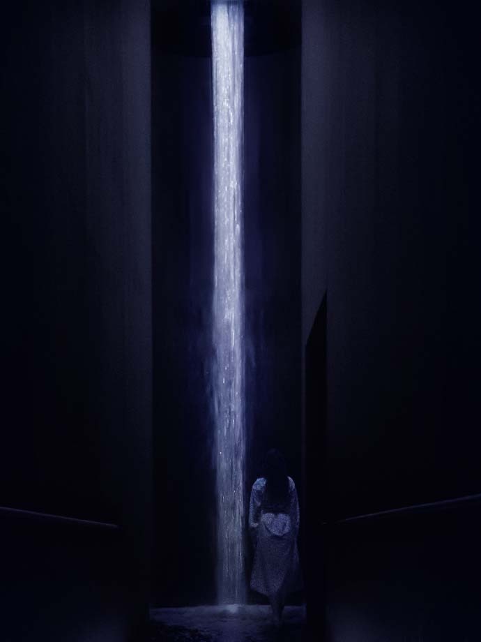 A lady in white standing in the middle of a dark room staring at a waterfall of light